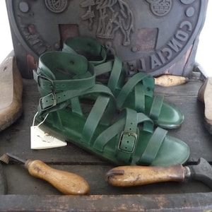 K&ouml;nnte beinhalten: Ein Paar gr&uuml;ne Ledersandalen mit mehreren Riemen und Schnallen. Die Sandalen befinden sich auf einer Holzoberfl&auml;che.