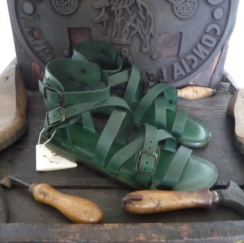 K&ouml;nnte beinhalten: Ein Paar gr&uuml;ne Ledersandalen mit mehreren Riemen und Schnallen. Die Sandalen befinden sich auf einer Holzoberfl&auml;che.