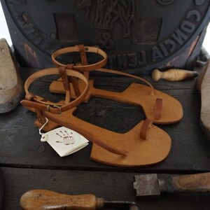 May include: A pair of brown leather sandals with a simple design. The sandals have a strap that goes around the ankle and a toe strap. The sandals are handmade and have a tag that says "Handmade".