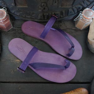 May include: Two pairs of purple leather sandals with a toe loop and ankle strap. The sandals have a simple design and are made of soft leather.