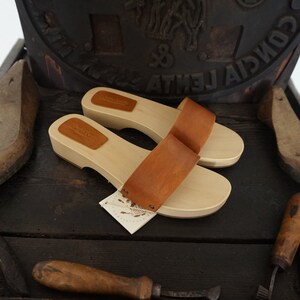 May include: A pair of wooden clogs with light brown leather straps. The clogs have a natural wood finish and are displayed on a dark wooden surface. A tag is attached to one of the clogs.