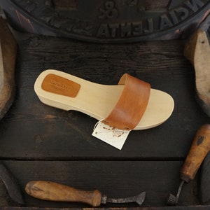 May include: A wooden sandal with a light brown sole and a wide, medium-brown leather strap. The sandal has a small leather patch with text on the top. A tag is attached to the sandal. The sandal is displayed on a dark wooden surface.