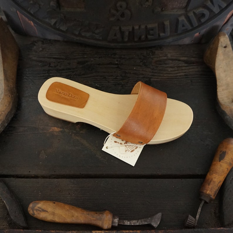 May include: A wooden sandal with a light brown sole and a wide, medium-brown leather strap. The sandal has a small leather patch with text on the top. A tag is attached to the sandal. The sandal is displayed on a dark wooden surface.