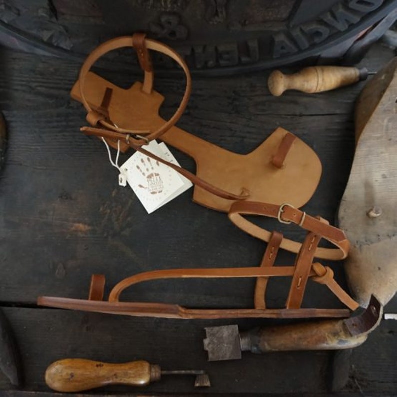 May include: A pair of brown leather sandals with a strap across the foot and ankle. The sandals are handmade and have a simple design. The sandals are on a wooden surface with a small tool and a piece of leather.