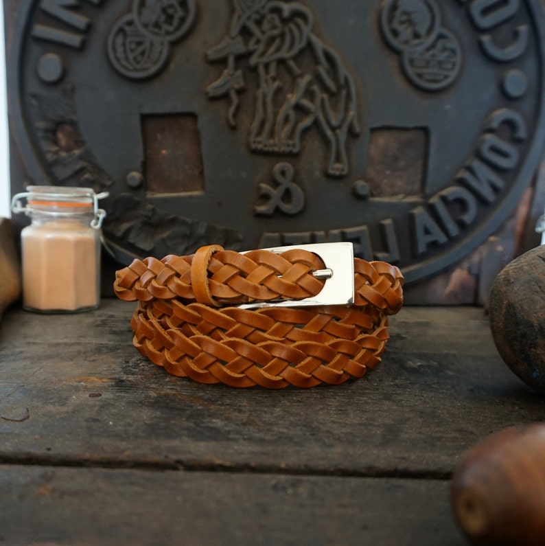May include: A brown leather braided belt with a silver buckle. The belt is folded and lying on a wooden surface.