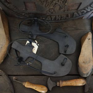 May include: Two black leather sandals with straps and buckles, along with wooden shoe lasts and leather working tools on a wooden surface.