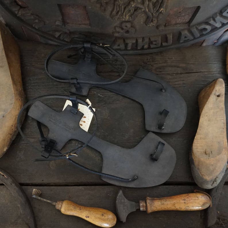 May include: Two black leather sandals with straps and buckles, along with wooden shoe lasts and leather working tools on a wooden surface.
