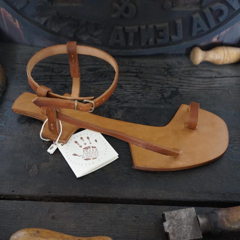 May include: A brown leather sandal with a strap that goes around the ankle and a toe loop. The sandal is handmade and has a label that says "Petite  Couture  Handmade in Greece".