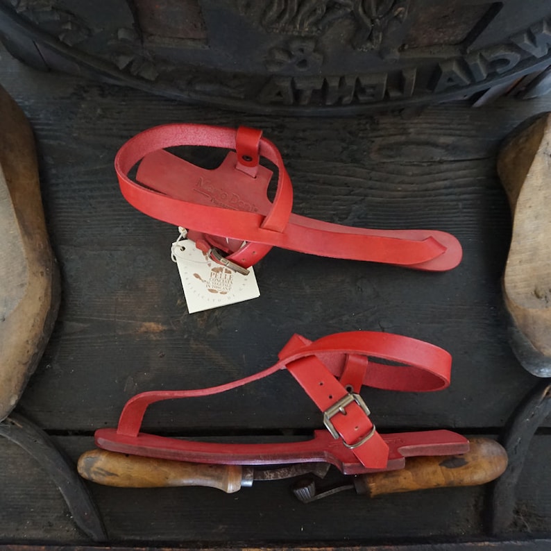 May include: A pair of red leather sandals with a buckle strap. The sandals are handmade and have a wooden sole.