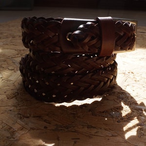 Puede incluir: Un cintur&oacute;n de cuero marr&oacute;n con un dise&ntilde;o trenzado. El cintur&oacute;n tiene una hebilla plateada y un nudo tejido en el extremo.
