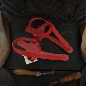 May include: A pair of red leather sandals with a simple design. The sandals have a strap that goes around the ankle and a toe strap. The sandals are made of leather and have a leather sole.