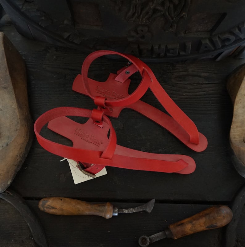 May include: A pair of red leather sandals with a simple design. The sandals have a strap that goes around the ankle and a toe strap. The sandals are made of leather and have a leather sole.