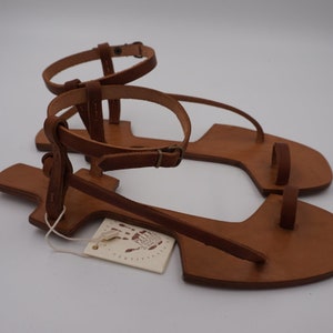 May include: A pair of brown leather sandals with a toe loop and ankle strap. The sandals have a simple design and are made of high-quality leather. The sandals are handmade and feature a tag that says "Handmade with Love".