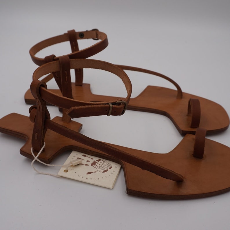 May include: A pair of brown leather sandals with a toe loop and ankle strap. The sandals have a simple design and are made of high-quality leather. The sandals are handmade and feature a tag that says "Handmade with Love".