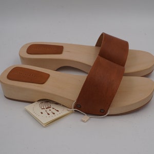 May include: A pair of wooden clogs with light brown wooden soles and brown leather straps. The clogs have a tag attached with the text "MARIO DONI" and a handprint design. The shoes are new and unworn.
