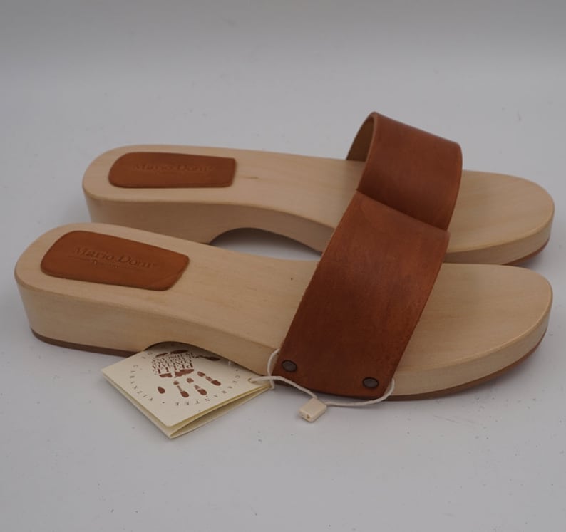 May include: A pair of wooden clogs with light brown wooden soles and brown leather straps. The clogs have a tag attached with the text "MARIO DONI" and a handprint design. The shoes are new and unworn.