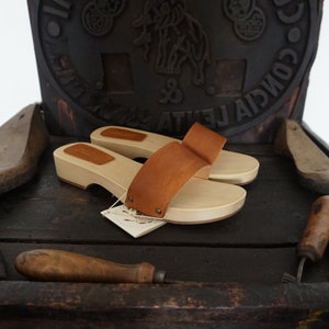 May include: A pair of wooden clogs with light brown leather straps. The clogs have a natural wood finish and are displayed on a dark wooden surface. A tag is attached to one clog. The background includes tools and a decorative element.