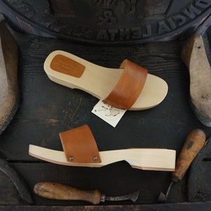 May include: Two wooden clogs with brown leather straps and a light wood base. The clogs are displayed with shoemaking tools and a wooden shoe form. The clogs have a tag that says "Mano Doni".