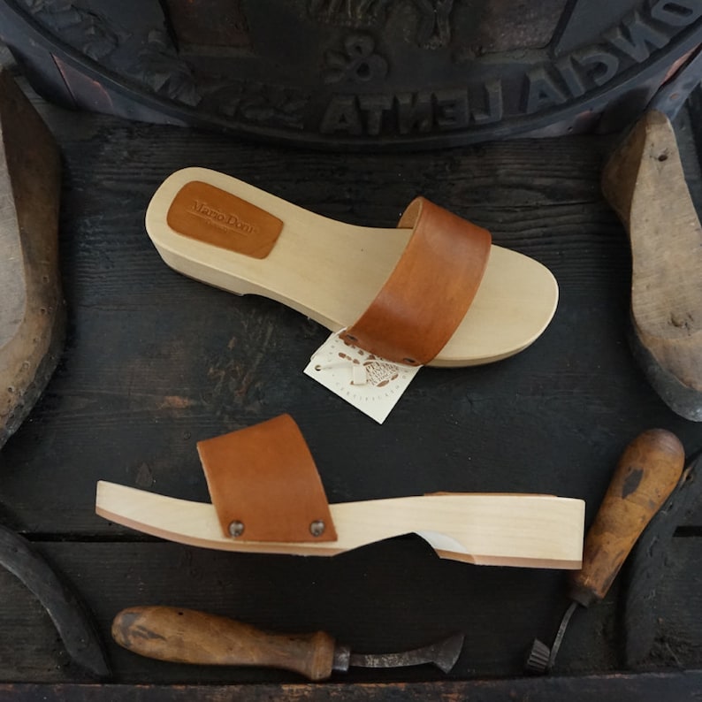 May include: Two wooden clogs with brown leather straps and a light wood base. The clogs are displayed with shoemaking tools and a wooden shoe form. The clogs have a tag that says "Mano Doni".