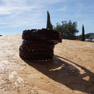 Puede incluir: Un cintur&oacute;n de cuero marr&oacute;n con un dise&ntilde;o trenzado y una hebilla plateada. El cintur&oacute;n est&aacute; doblado por la mitad y est&aacute; sobre una superficie de madera.