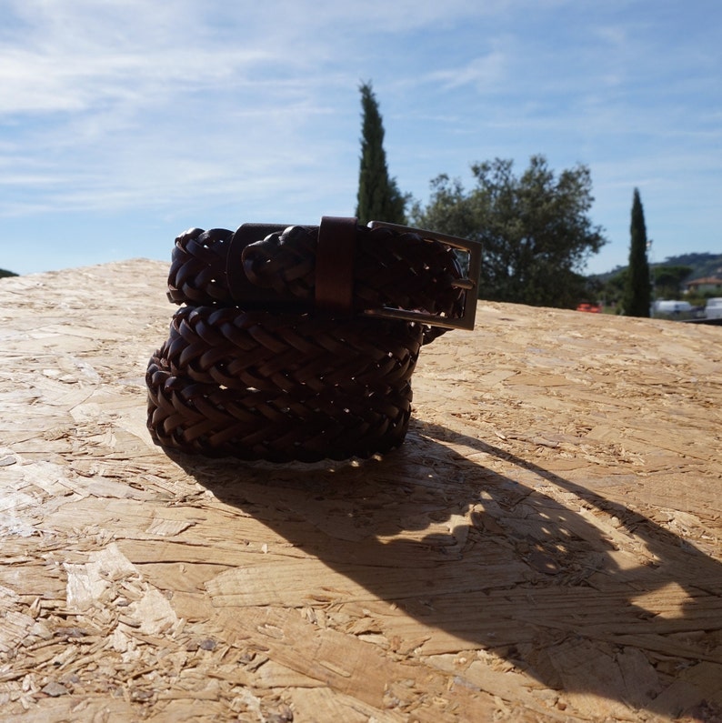 Puede incluir: Un cintur&oacute;n de cuero marr&oacute;n con un dise&ntilde;o trenzado y una hebilla plateada. El cintur&oacute;n est&aacute; doblado por la mitad y est&aacute; sobre una superficie de madera.