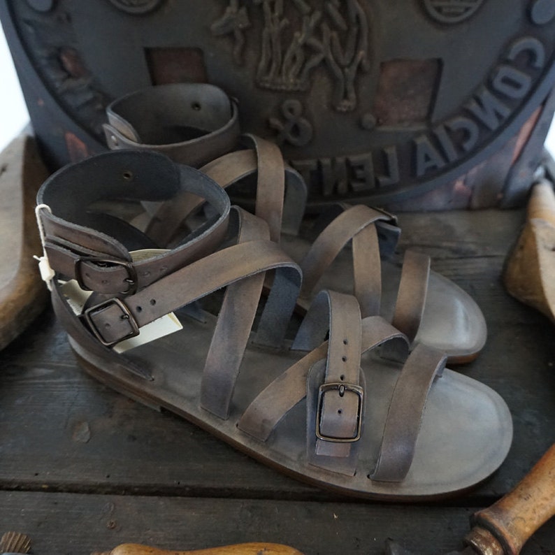 K&ouml;nnte beinhalten: Ein Paar braune Ledersandalen mit mehreren Riemen und Schnallen. Die Sandalen haben einen abgenutzten, verwitterten Look und sind aus dickem, strapazierf&auml;higem Leder gefertigt.