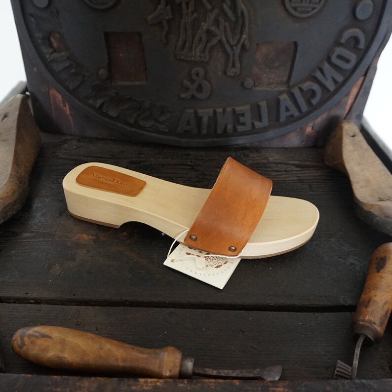 May include: A wooden clog with a light brown sole and a wide, tan leather strap. The clog has a small, rectangular leather patch on the top. A white tag is attached to the clog. The clog is displayed on a dark wooden surface.