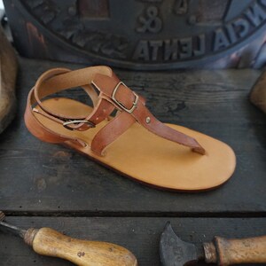Pu&ograve; includere: Un paio di sandali in pelle marrone con cinturino con fibbia e cinturino per le dita. I sandali hanno un design semplice e sono realizzati in pelle di alta qualit&agrave;.