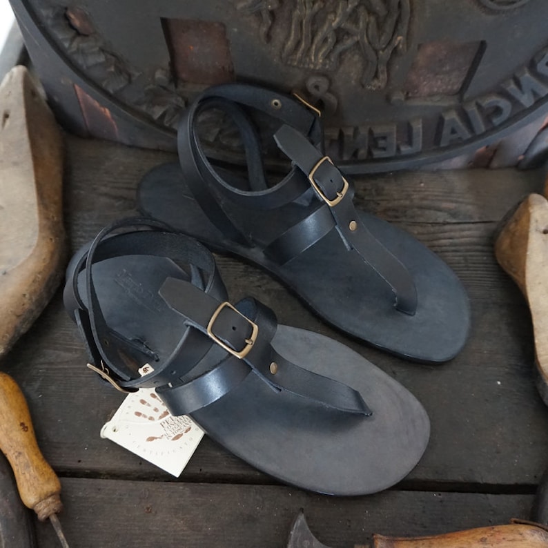 Pu&ograve; includere: Un paio di sandali in pelle nera con chiusura a fibbia e cinturino a T. I sandali hanno un design semplice e sono realizzati in pelle di alta qualit&agrave;.