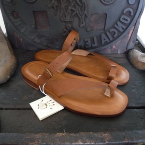 May include: A pair of brown leather sandals with a toe thong and ankle strap. The sandals have a simple design and are made of high-quality leather.