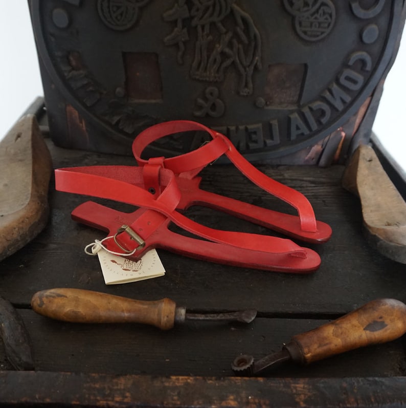 May include: Red leather sandals with a buckle fastening. The sandals are handmade and have a simple design. The sandals are on a wooden surface with leather working tools.
