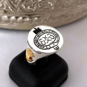 May include: A silver ring with a gold accent. The ring features a crest with the text "SOIT QUI MAL Y PENSE" and "HONI SOIT QUI MAL Y PENSE". The ring also has the text "GRENADIER GUARDS" on the side.