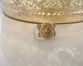 Puede incluir: Un anillo de plata y oro con un símbolo masónico en la parte superior. El símbolo es una brújula y una escuadra con una 'G' en el centro.