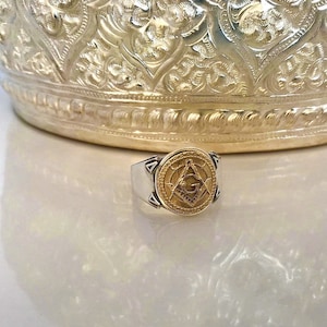 Puede incluir: Un anillo de plata y oro con un símbolo masónico en la parte superior. El símbolo es una brújula y una escuadra con una 'G' en el centro.