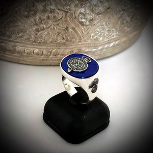 Puede incluir: Un anillo de plata con una piedra azul y un emblema de plata en la parte superior. El emblema tiene una corona y el número 288.