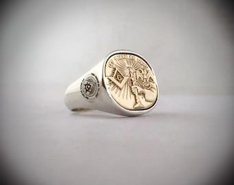 May include: A silver ring with a gold Masonic symbol. The symbol features a man holding a torch and a square and compass. The text "Let There Be Light" is inscribed around the symbol.