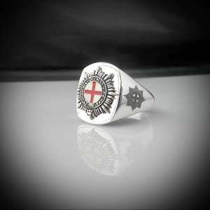 May include: A silver signet ring with a raised design of a cross and a starburst. The ring is square shaped and has a smooth, polished finish.