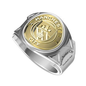 Puede incluir: Un anillo de plata y oro con el logotipo del Manchester City Football Club. El anillo tiene un diseño en relieve y las palabras "Manchester City Football Club" están grabadas en el lateral.