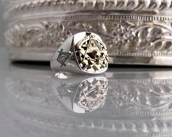 May include: A silver ring with a gold Masonic symbol and skull design. The ring is set against a silver background.