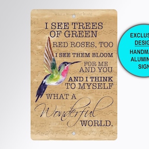 May include: A rectangular aluminum sign with a tan background featuring musical notation. It displays a watercolor hummingbird and the text: I SEE TREES OF GREEN, RED ROSES, TOO, I SEE THEM BLOOM FOR ME AND YOU, AND I THINK TO MYSELF WHAT A WONDERFUL WORLD.