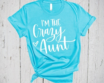 May include: A bright turquoise short-sleeve t-shirt featuring the white printed text I'M THE Crazy Aunt with a small heart icon. The shirt has a crew neckline and is tied in a knot at the bottom hem, displaying a casual and simple design.