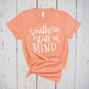 Puede incluir: Una camiseta de color melocotón con el texto "Southern State of Mind" en letras blancas.