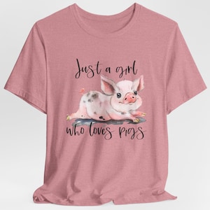 May include: A dusty rose t-shirt featuring a watercolor illustration of a pink piglet with black spots. The text "Just a girl who loves pigs" is written above and below the piglet in a handwritten font.