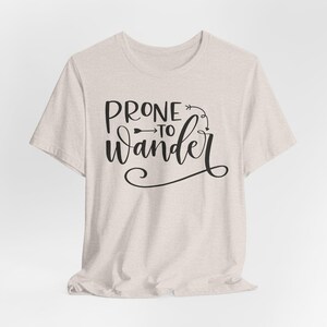 May include: A light beige t-shirt with the phrase "PRONE to Wander" in black script. The text is accompanied by an arrow and decorative flourishes, creating a casual and inviting design. The shirt is made of a soft, textured fabric.