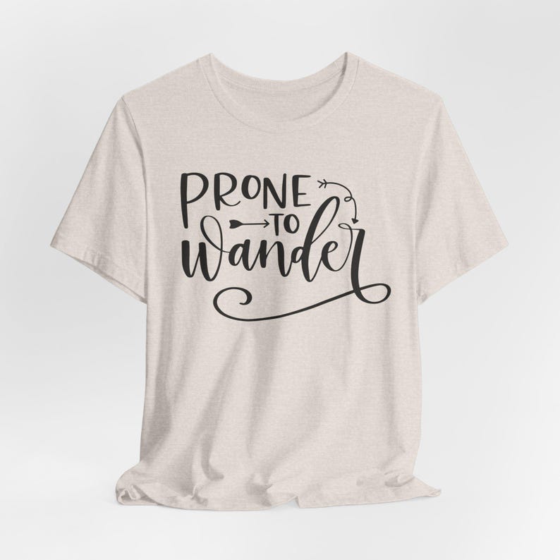 May include: A light beige t-shirt with the phrase "PRONE to Wander" in black script. The text is accompanied by an arrow and decorative flourishes, creating a casual and inviting design. The shirt is made of a soft, textured fabric.