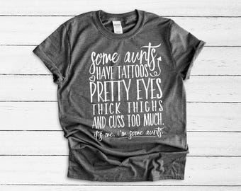 May include: A dark heather grey short-sleeve t-shirt with white text that reads: Some aunts have tattoos, pretty eyes, thick thighs and cuss too much. It's me. I'm some aunts. The shirt features a crew neckline and a standard fit.