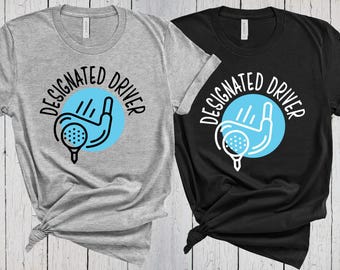 May include: A heather grey t-shirt and a black t-shirt, each featuring a graphic of a golf club hitting a ball on a tee inside a light blue circle. The text "DESIGNATED DRIVER" is printed in a circular arc above the golf graphic.