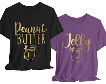 May include: A black t-shirt with gold text reading Peanut BUTTER and a gold jar graphic, alongside a purple t-shirt with gold text reading Jelly and a gold jar graphic. Both shirts feature short sleeves and a crew neckline design.