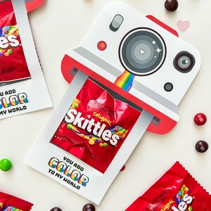 May include: A white and red cardboard camera with a rainbow and a small bag of Skittles candy inside. The text "You add color to my world" is printed on the front of the camera.