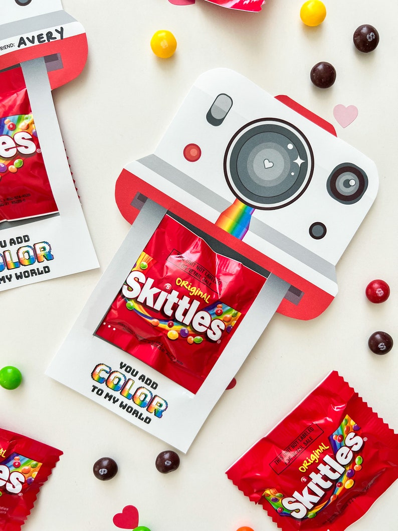 May include: A white and red cardboard camera with a rainbow and a small bag of Skittles candy inside. The text "You add color to my world" is printed on the front of the camera.